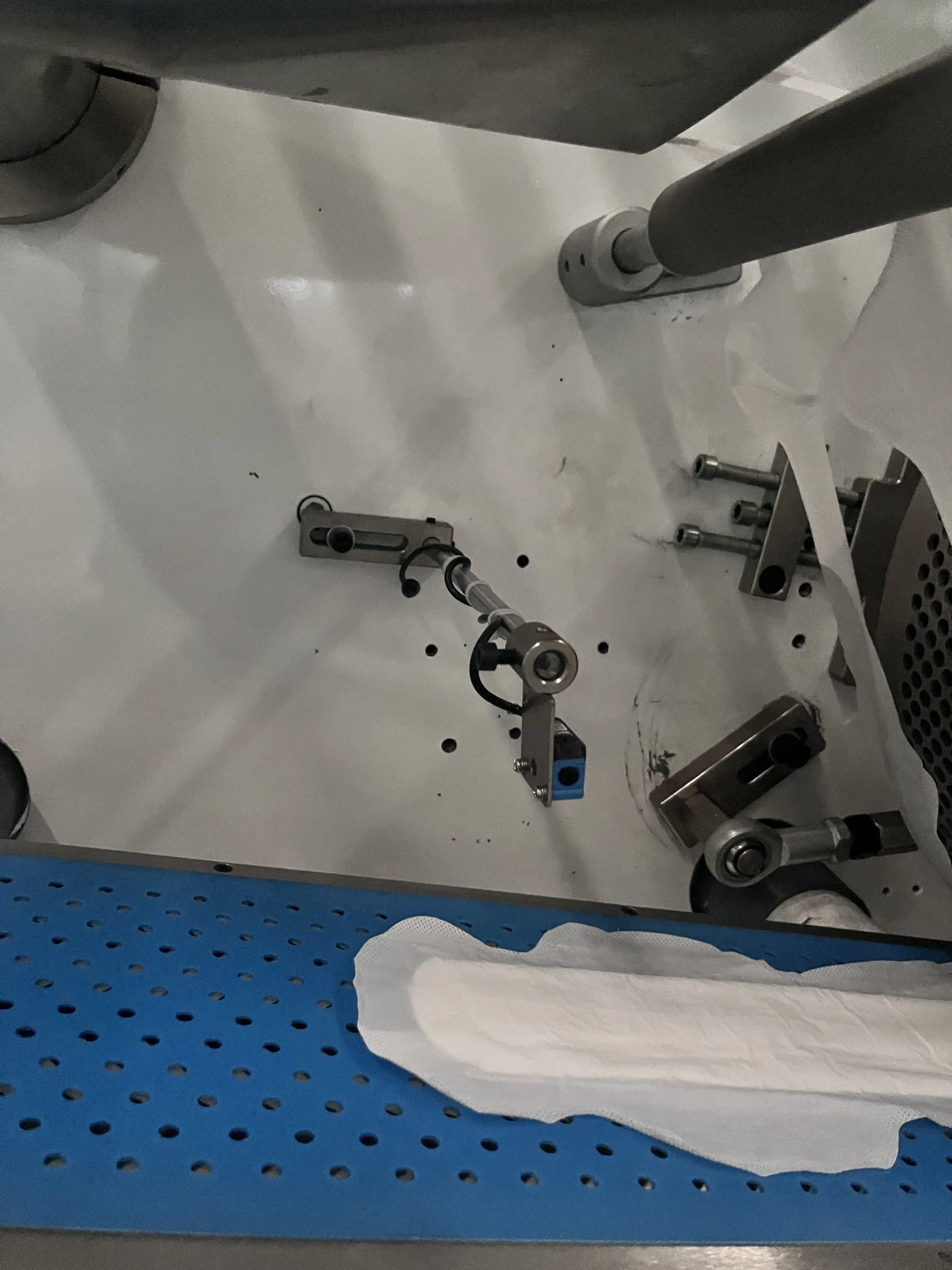 Inline sensor array over a blue perforated conveyor on a Welldone sanitary napkin line — validates every pad for length, thickness, and join integrity as part of the 30-day production audit trail